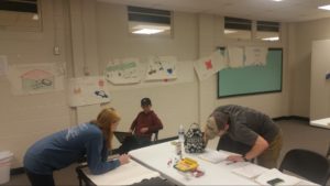 Three students work around a table; one stands over a spiral notebook, reading text; another leans over white paper, sketching; a third sits in a chair, using a laptop. Storyboard appears on wall behind them.