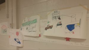 Various large pieces of white paper taped to cinder-block wall; marker-drawn sketches of stick figures (murder victims), hotel rooms, gas stations, and police vehicles